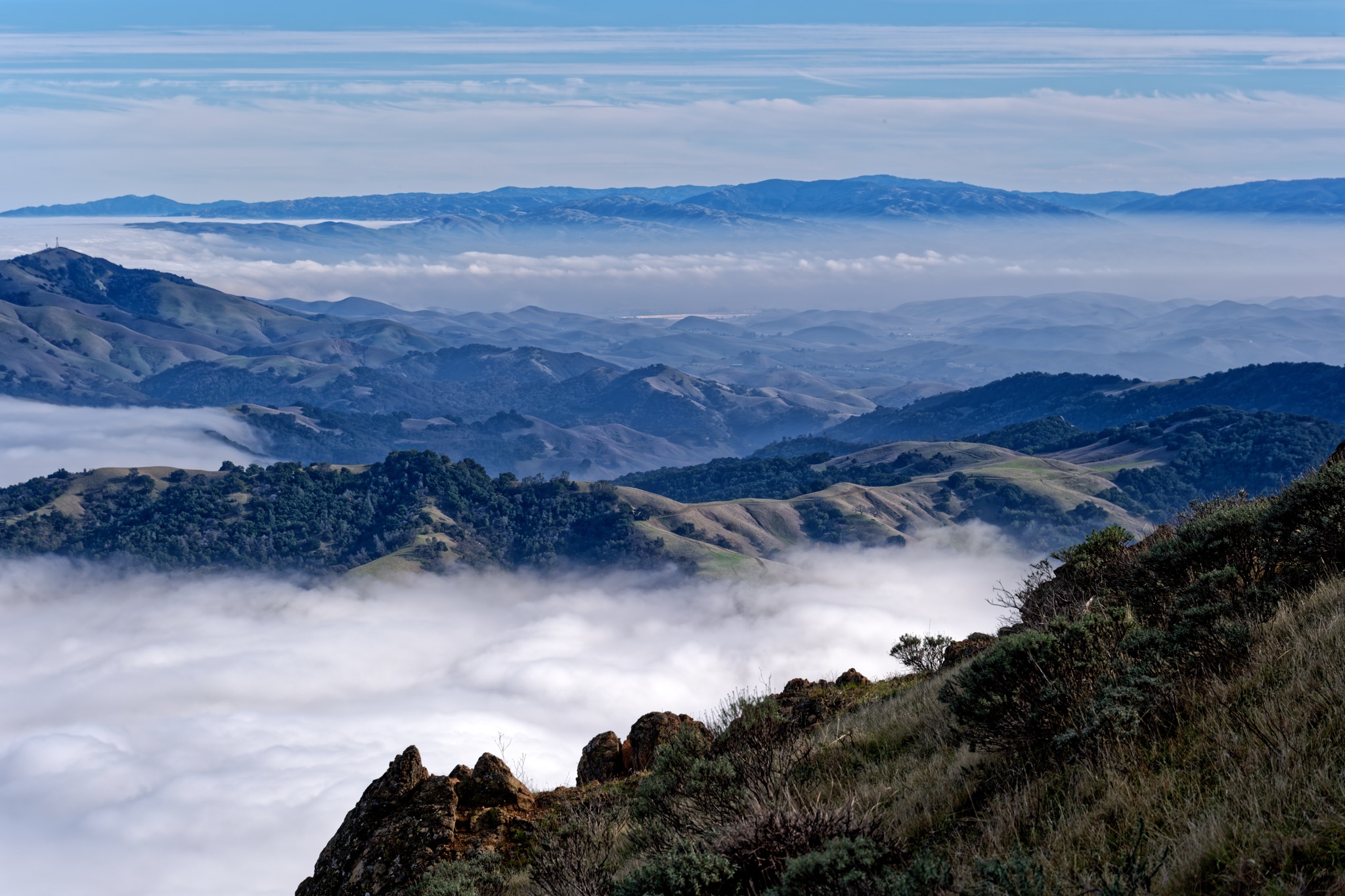 Rugged, grassy hills and rocky outcrops rise above a thick layer of white fog that fills the valleys below.