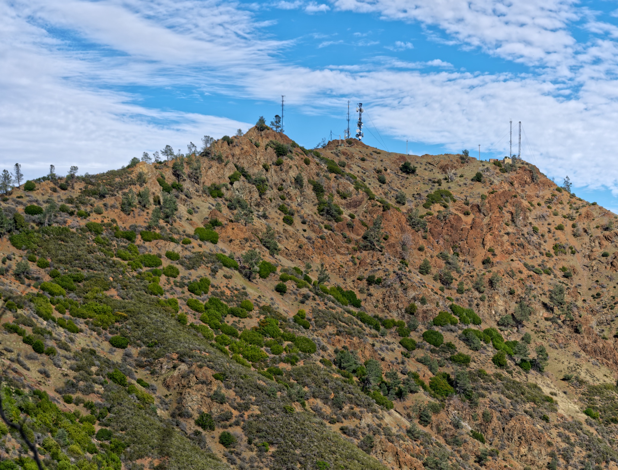 Rugged, reddish-brown slopes of Mt Diablo's North Peak Trail rise under a blue sky, dotted with green shrubs and topped by several tall communication towers.