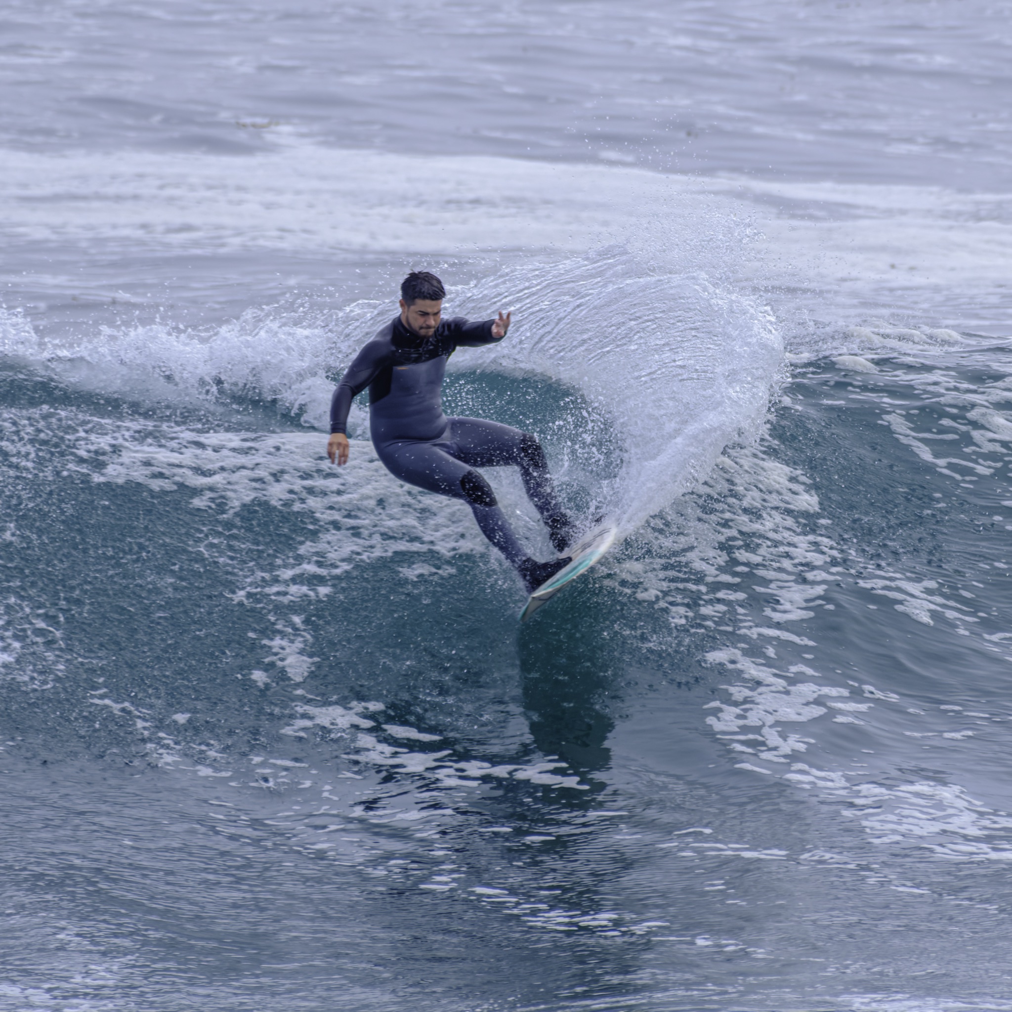 A man in a black wetsuit crouches on a surfboard, carving a sharp turn through a breaking wave and spraying a fan of white water.