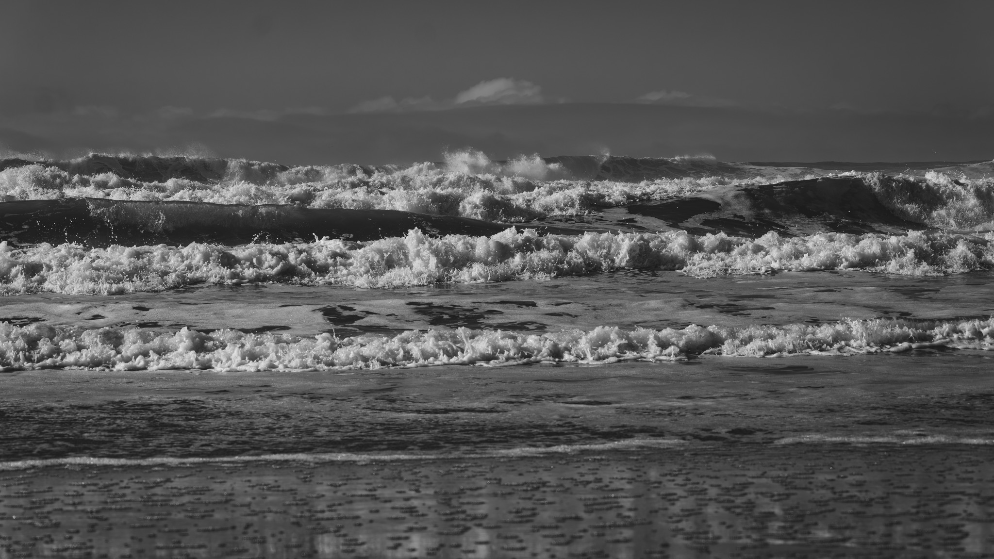 Black and white view of breaking ocean waves and white foam rolling onto the wet sand at Pescadero Beach under a cloudy sky.
