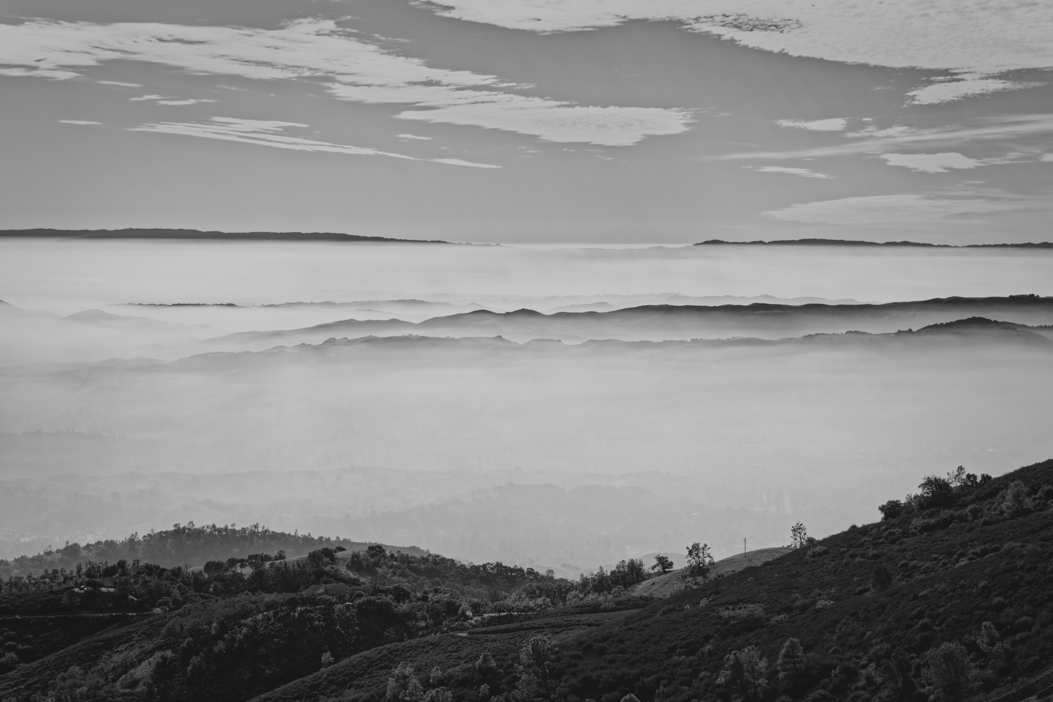 A black and white view of layered mountain ridges fading into a thick sea of fog beneath a cloudy sky.