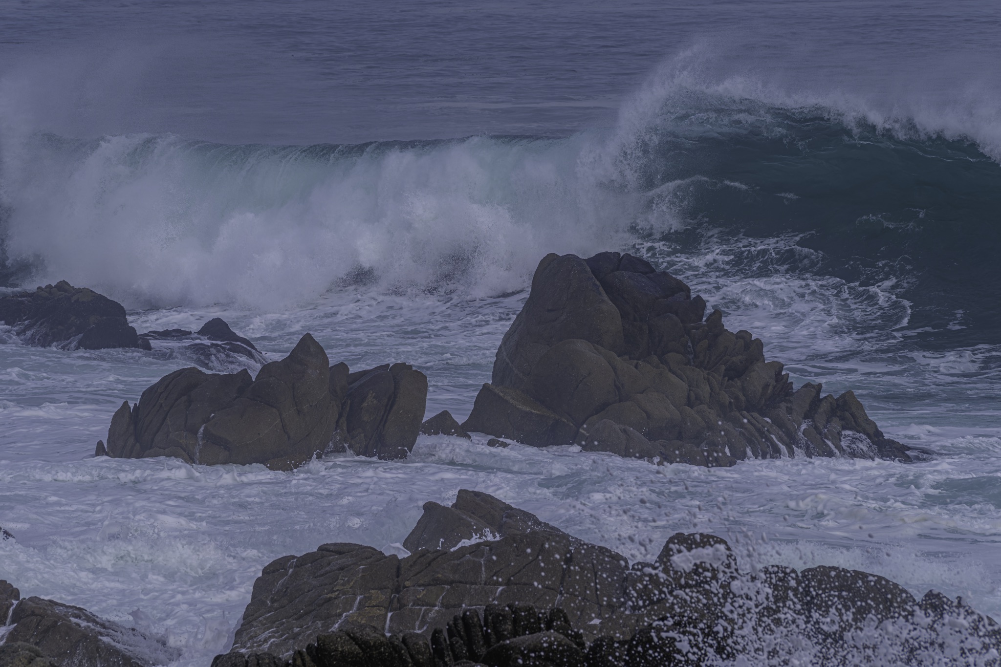 Large ocean waves crash against dark, jagged rocks along the Monterey waterfront, sending white spray high into the air.
