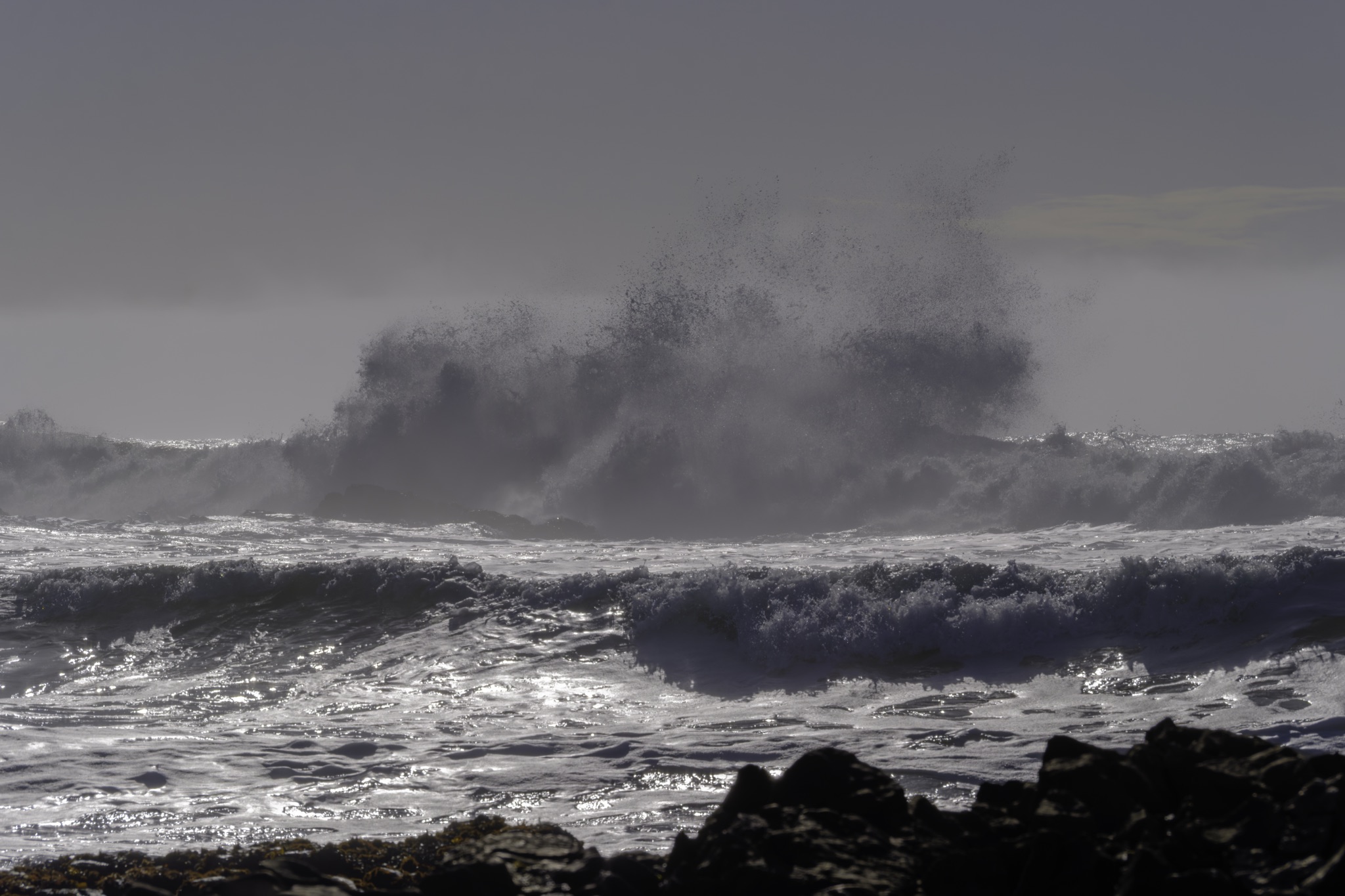 A large wave crashes against dark rocks at Pescadero Beach, sending a dramatic spray of water high into the air under a hazy sky.