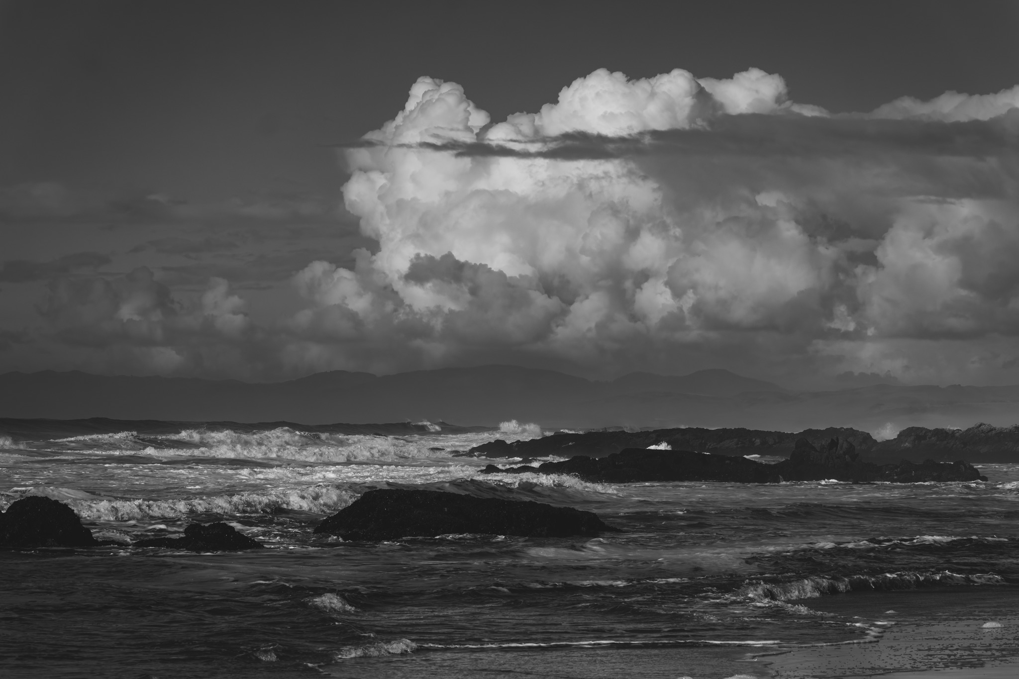 Black and white view of waves crashing on dark rocks at Pescadero Beach, with a large cumulus cloud above distant mountains.