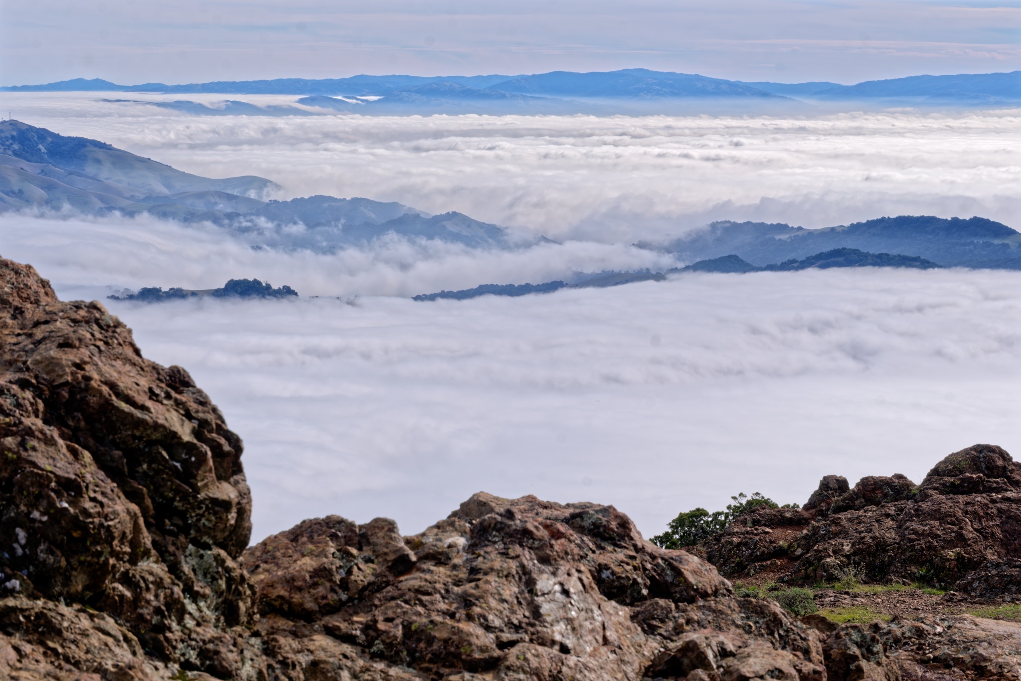 Rugged brown rocks frame a view of a thick sea of clouds filling the valleys, with distant blue mountain ridges rising above the mist.