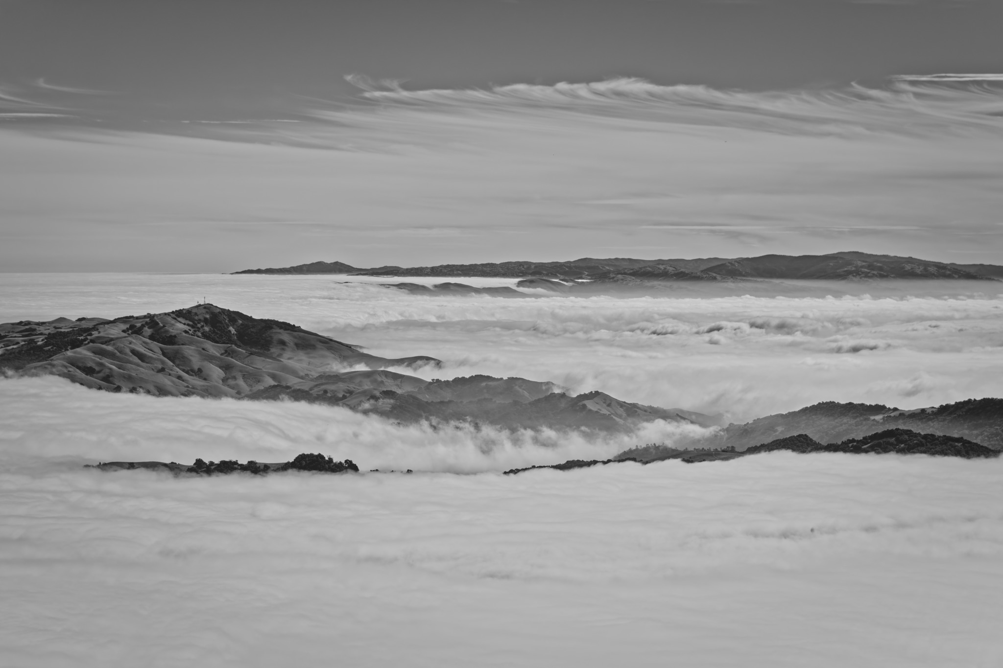 Black and white view of rolling hills emerging from a thick sea of clouds beneath a sky with wispy cirrus clouds.