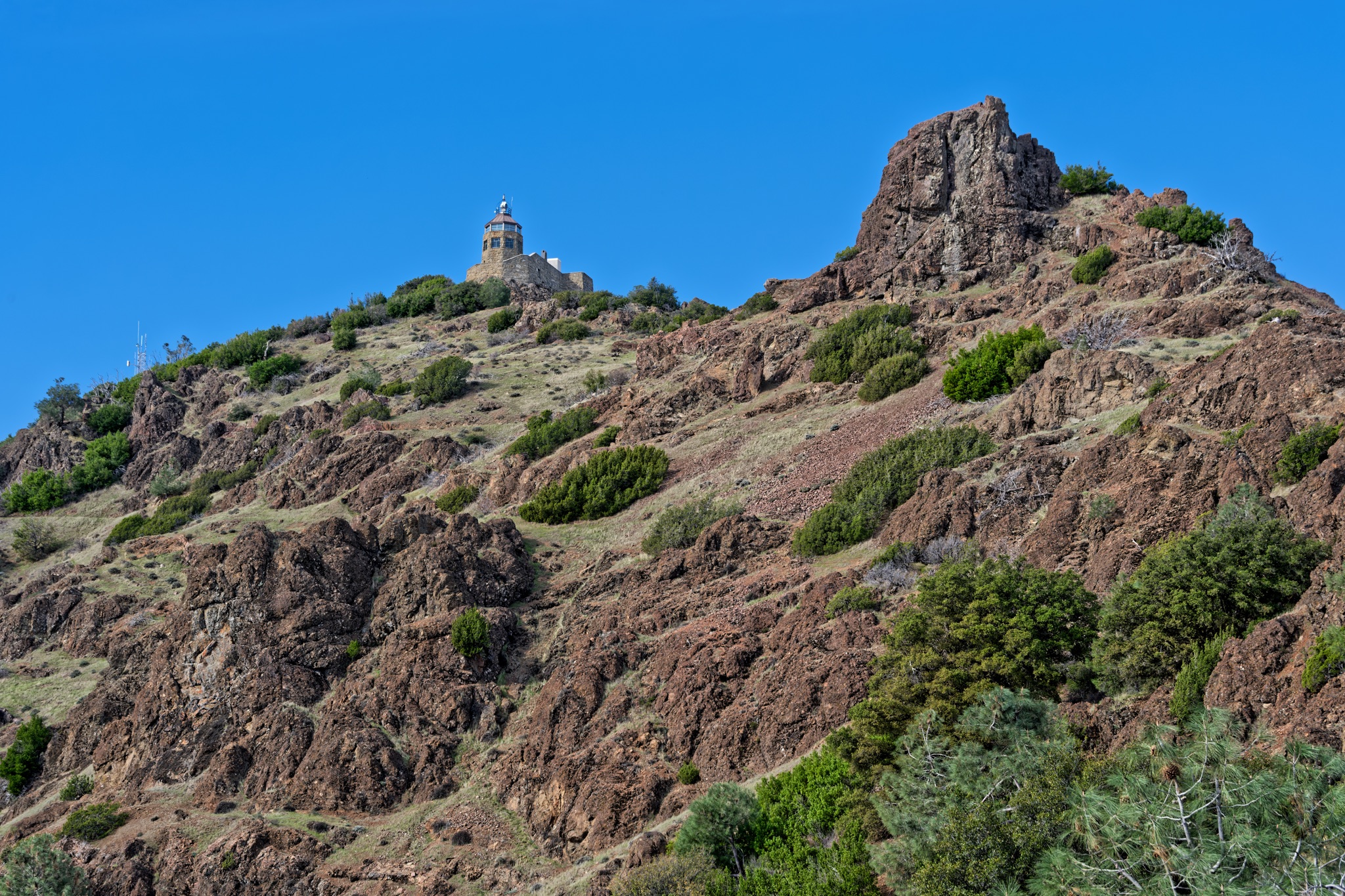 A stone lighthouse with a red roof sits atop a rugged, rocky hillside dotted with green shrubs under a clear blue sky.