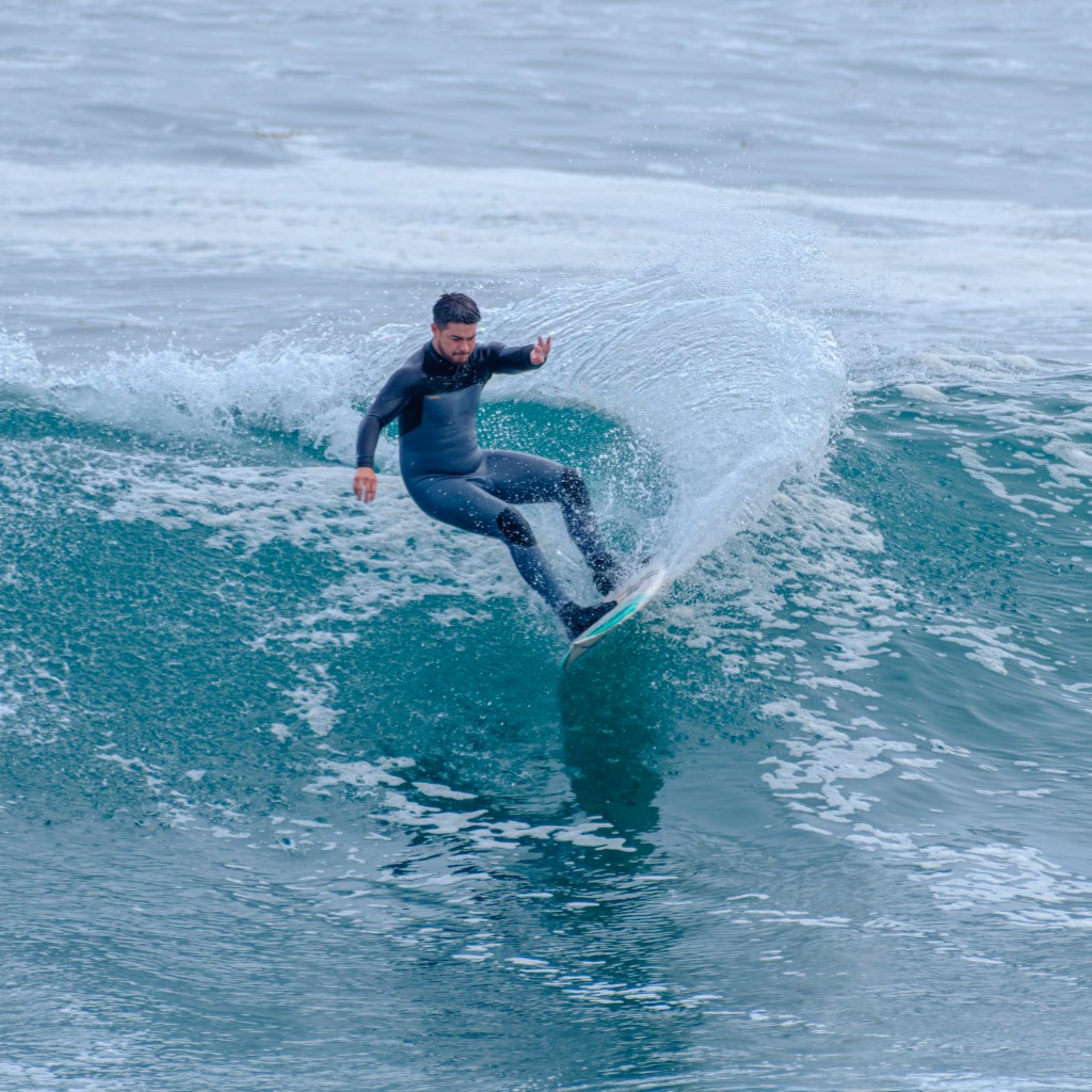 A man in a black wetsuit rides a surfboard along the face of a breaking wave, spraying water as he carves through the ocean.