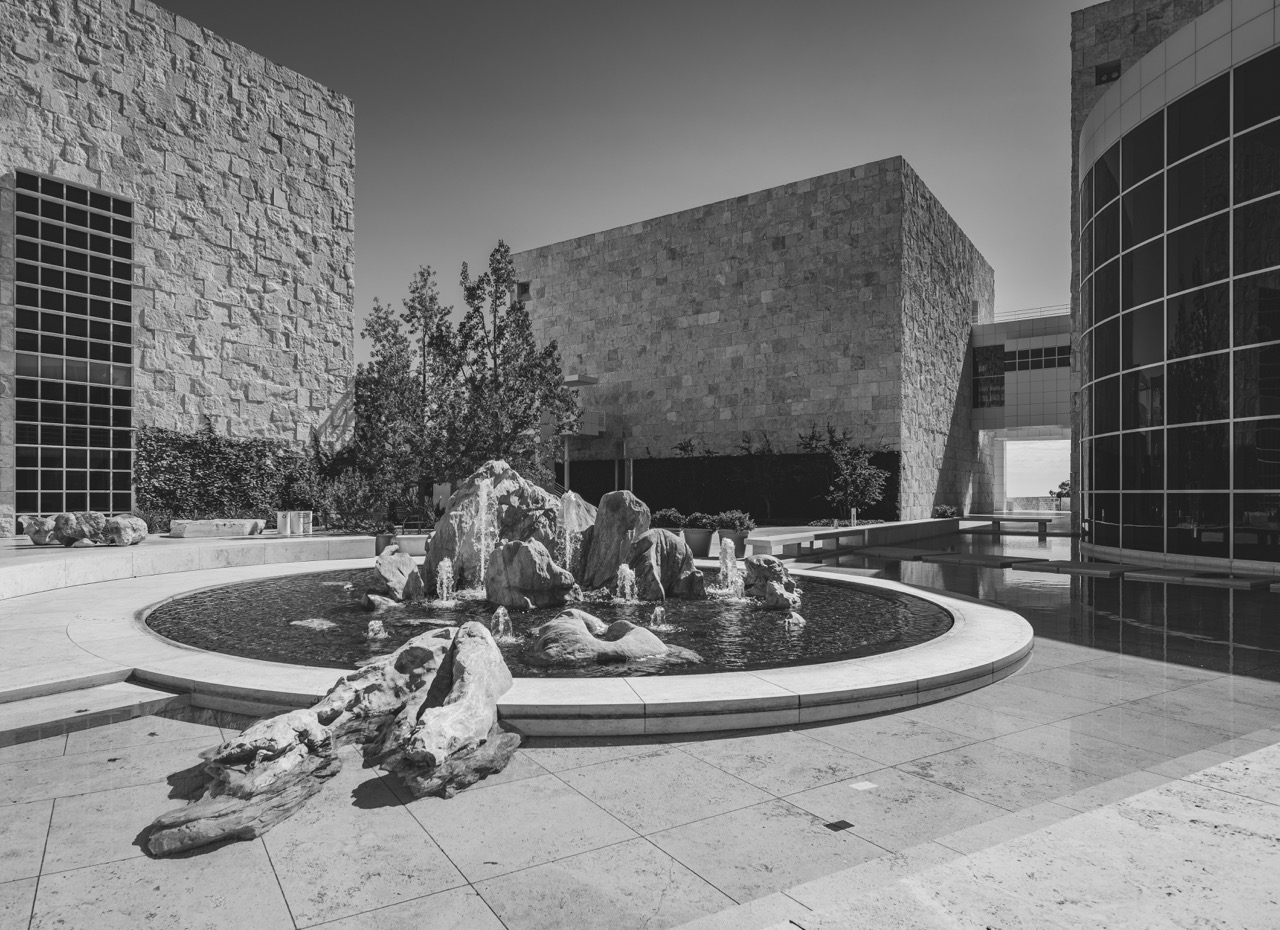 A circular fountain with rocks and water jets sits on a stone plaza at the Getty Center, surrounded by tall stone buildings and a curved glass wall.