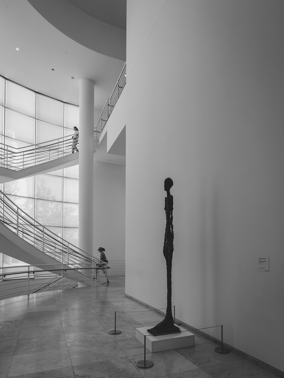 A tall, thin, dark sculpture of a human figure stands on a pedestal in a bright, modern museum atrium with a sweeping spiral staircase.