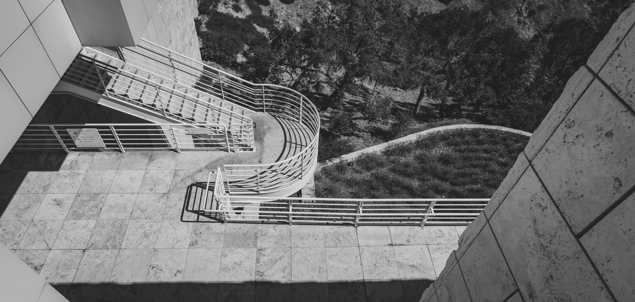 A high-angle view of a curved metal staircase and railings on a stone terrace at the Getty Center, with trees and a hillside visible below.