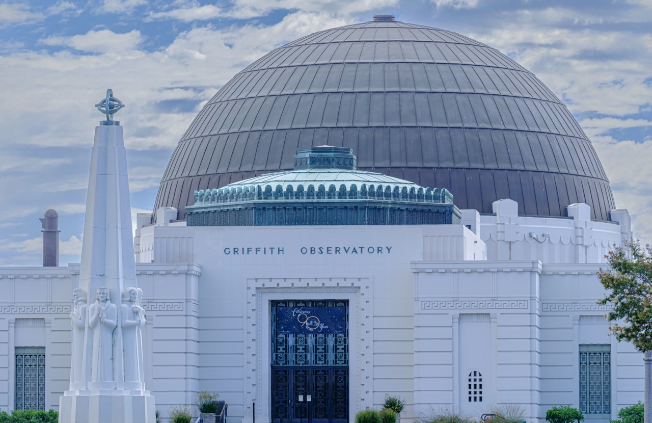 The white facade of Griffith Observatory in Los Angeles features a large dark dome and a tall white obelisk topped with a celestial armillary sphere.