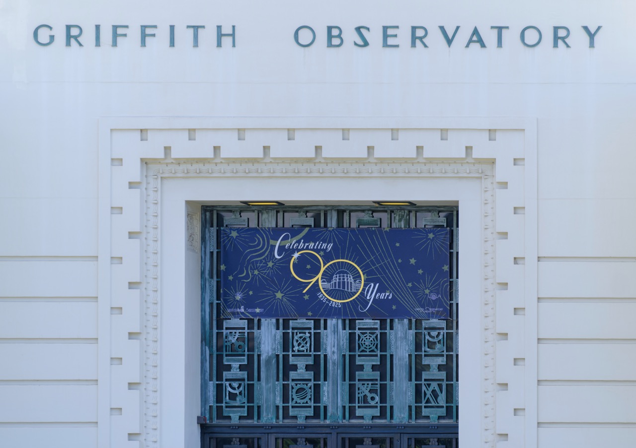 The white facade of Griffith Observatory in Los Angeles, California, with a blue banner reading "Celebrating 90 Years" above its ornate metal gate.