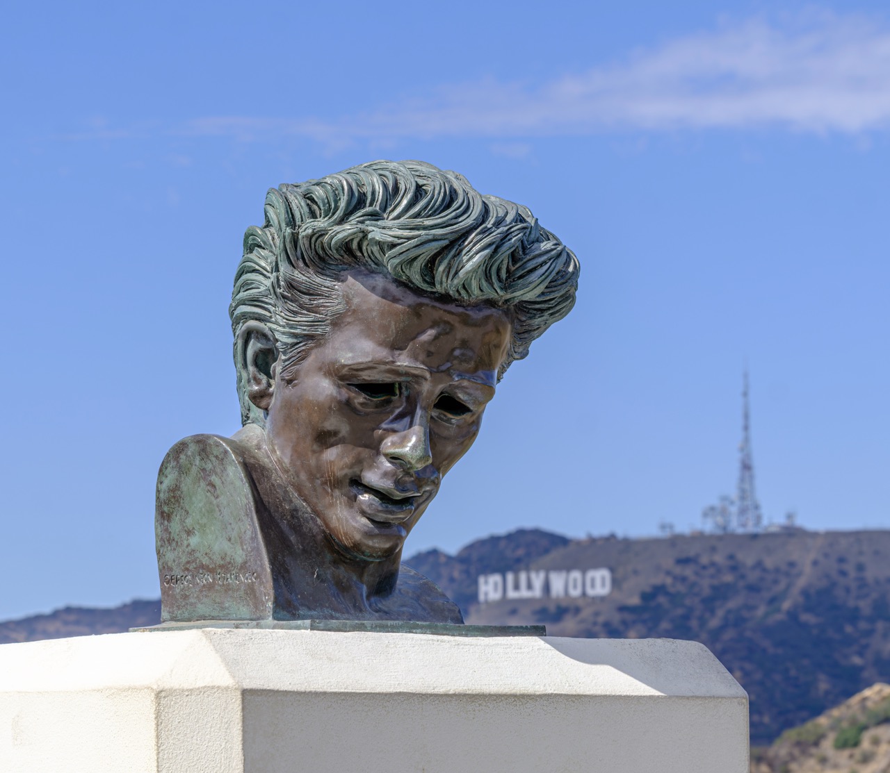 A bronze bust of James Dean with a green patina sits on a white pedestal, with the Hollywood sign visible on a hill in the background.