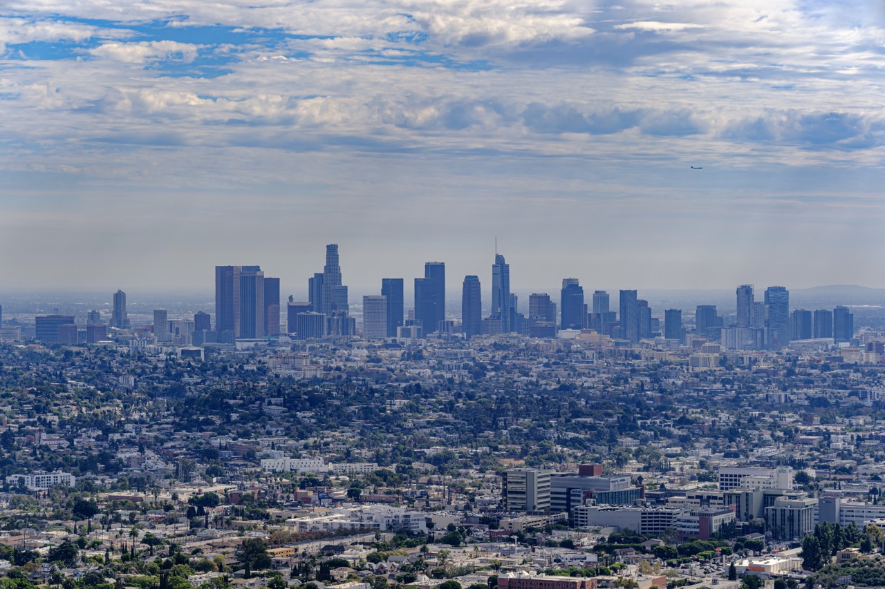 Aerial view of Los Angeles skyline with downtown skyscrapers rising above sprawling residential neighborhoods under a partly cloudy sky.