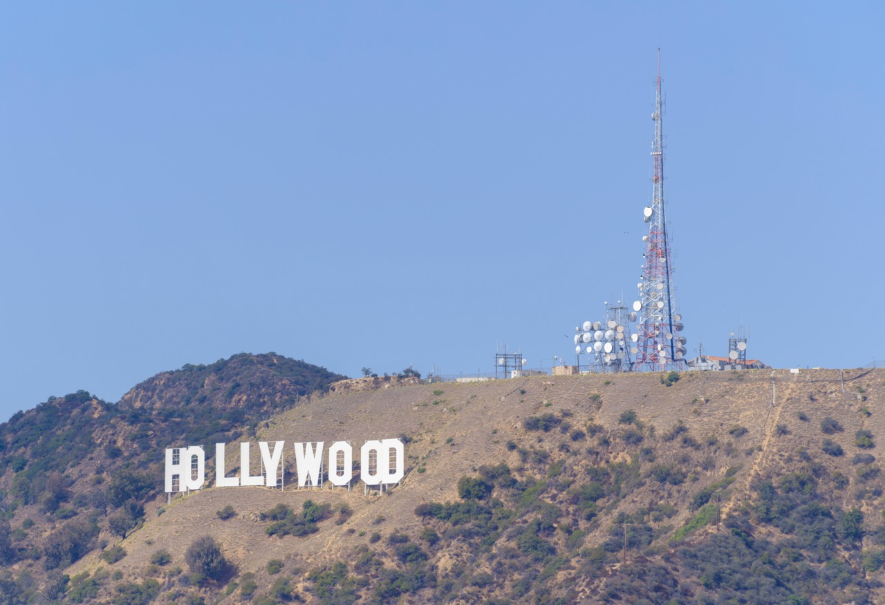 The white Hollywood sign sits on a dry, brush-covered hillside beneath a clear blue sky, with a tall red-and-white communications tower and satellite dishes on the ridge above.