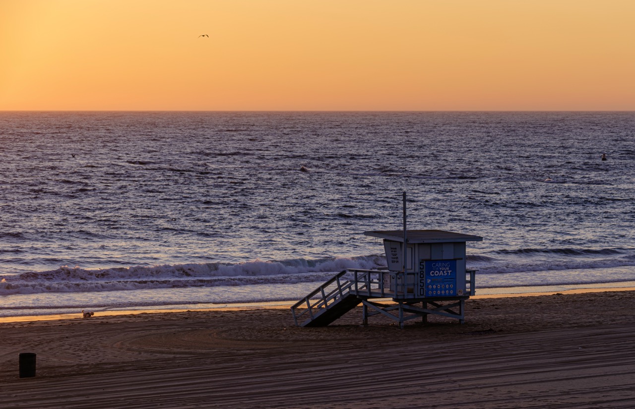 A lifeguard tower with a ramp sits on a sandy beach as the sun sets over the ocean, casting a warm orange glow across the sky.