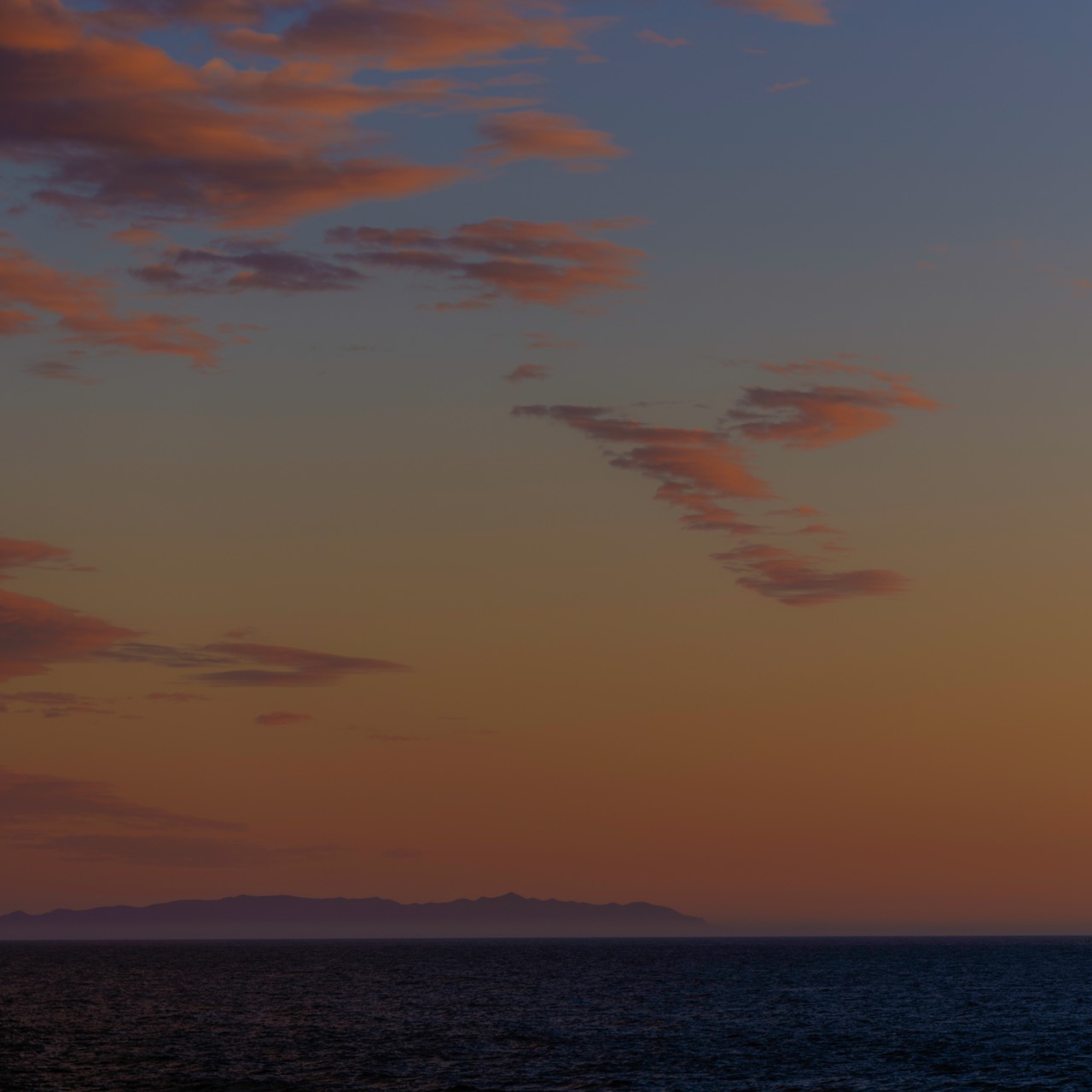 Orange and pink sunset clouds drift across a blue sky above a dark ocean and a distant mountain range.