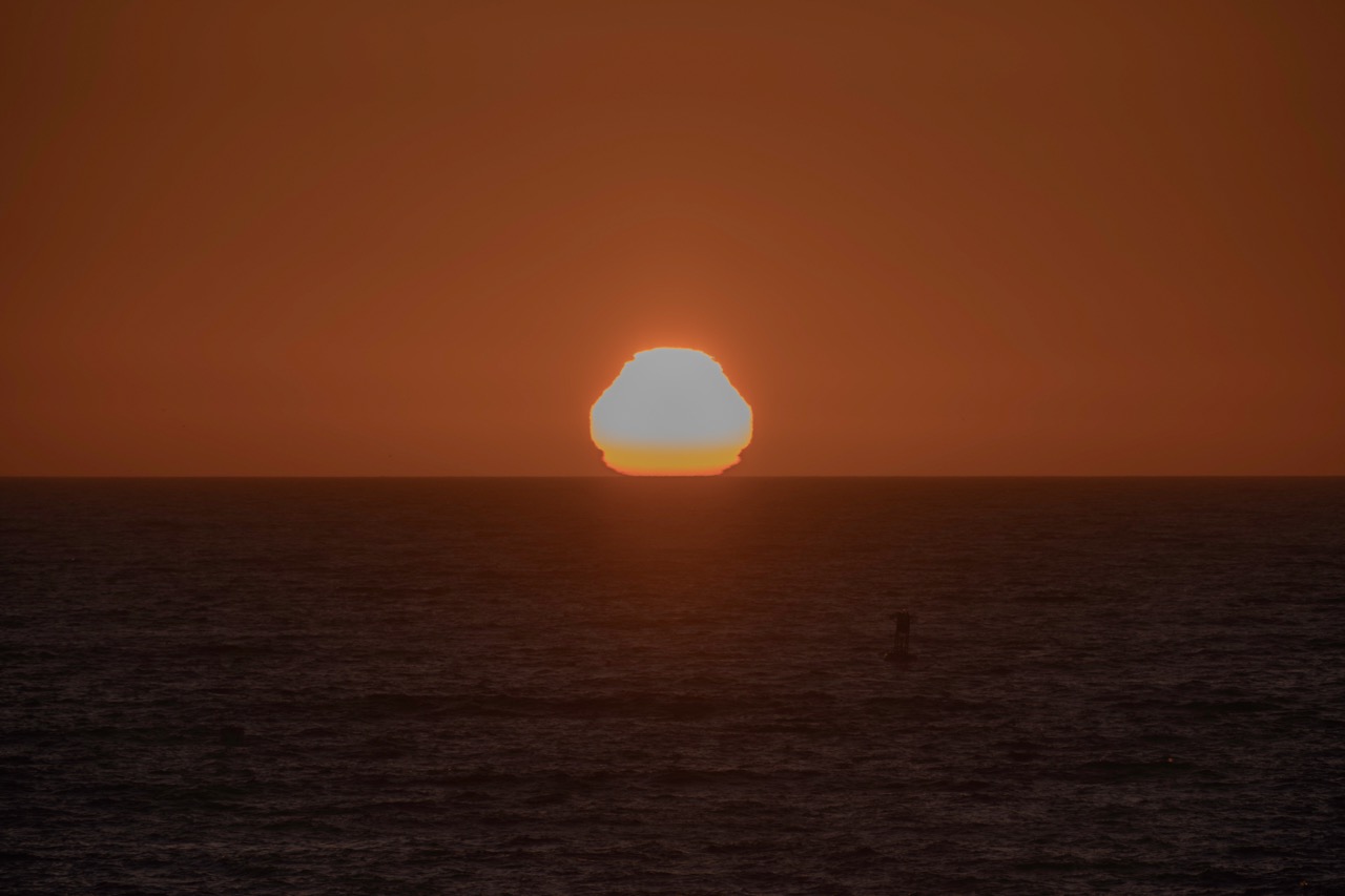 A bright sun sits low on the horizon over a dark, rippling sea, casting a deep orange glow across the sky.