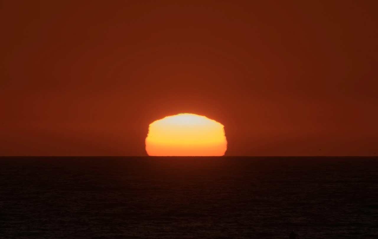 A bright orange sun sits low on the horizon over a dark ocean, casting a deep red glow across the sky.