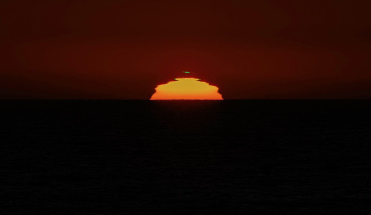 A bright orange sun sits low on the horizon, partially hidden by a dark cloud, over a black sea under a deep red sky.