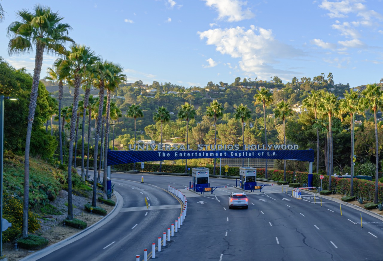 A car drives toward the Universal Studios Hollywood entrance gate, flanked by tall palm trees and green hills under a blue sky.