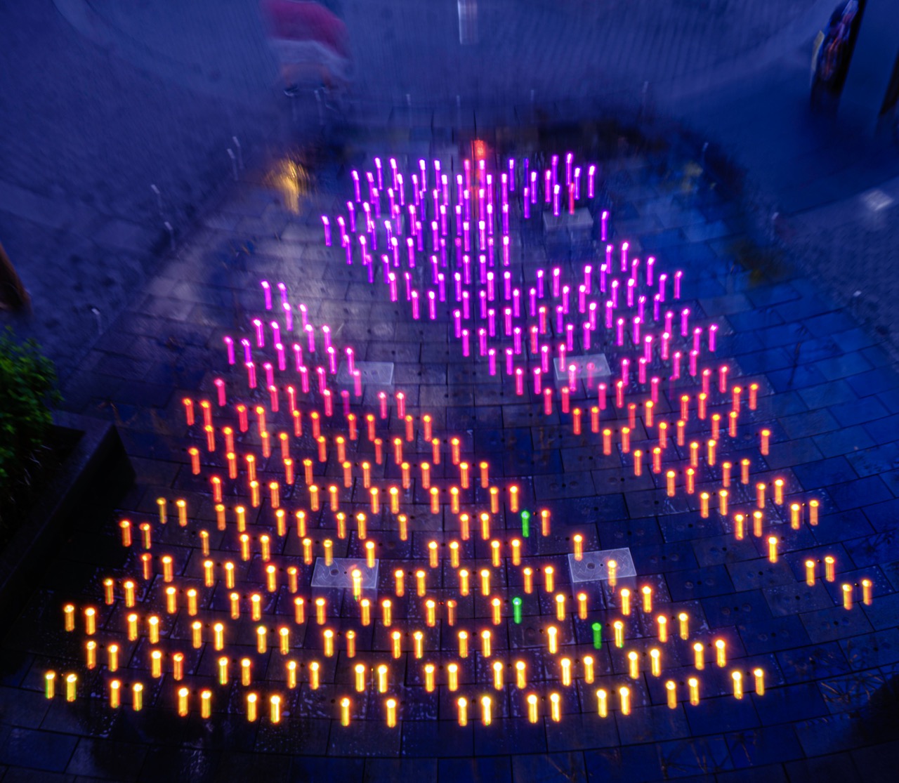 Hundreds of small candles in orange, pink, and green glow in a pattern on a dark stone plaza at night.